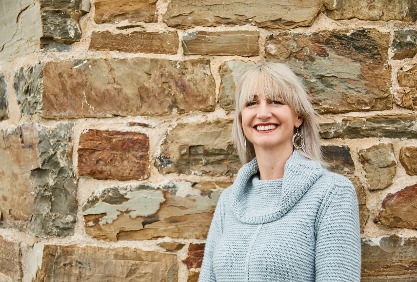 Sarah is wearing a blue jumper and leaning against a stone wall smiling at the camera.