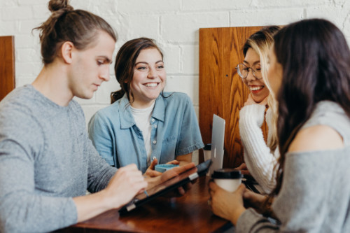 Three women and one man with a top knot, sitting in a cafe chatting over coffee